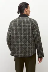 Campera capitoneada de corte recto con diseño a cuadros en tonos verde oliva y gris. Presenta cuello camisero de pana en color negro, cierre frontal con botones a presión y bolsillos laterales amplios con ribete negro.