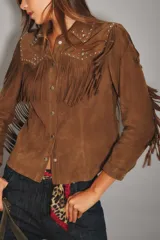 Camisa de gamuza color marrón con flecos en el frente, espalda y mangas. Presenta detalles de tachas metálicas en la parte superior delantera y cierre frontal con botones a presión.