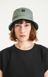 Gorro tipo bucket de color verde militar, con ala ancha y un pequeño parche negro con un logo blanco en el frente.