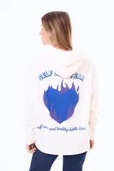 Buzo tipo canguro oversize de felpa color blanco, con capucha y bolsillo frontal. Presenta un diseño estampado en la espalda que incluye un corazón azul con llamas y la frase 'Self Love Team'.