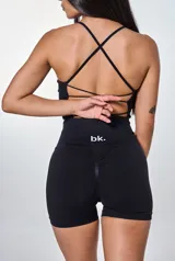 Conjunto deportivo de dos piezas en color negro, compuesto por un top con espalda descubierta y tiras cruzadas, y un short de tiro alto con efecto fruncido en la parte posterior. El short presenta el logo de la marca estampado en la cintura.