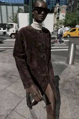 Abrigo marrón de gamuza con cuello redondo, manga larga, bolsillos delanteros y cierre frontal con botones.