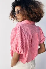Camisa corta de cuello solapa y manga corta abullonada acabada en elástico, color verde con estampado de cuadros vichy. Cuenta con un bolsillo de plastrón en el delantero, bajo con elástico y cierre frontal con botones.