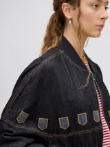 Campera tipo bomber confeccionada en denim de algodón color negro. Presenta un diseño holgado con cierre frontal metálico, bolsillos laterales con cierre y detalles decorativos en forma de escudos en la parte superior delantera.