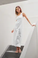 Vestido largo blanco de tirantes, corte recto y cuello cuadrado. Presenta un bordado vertical de flores en tonos grises en el lateral de la falda.