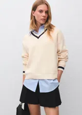 Jersey de punto grueso en color crema con cuello en V y detalles en contraste oscuro en el cuello y puños. Presenta un diseño recto de manga larga con una camisa de cuello camisero integrada en color celeste.