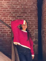 Buzo de tejido de punto con diseño a rayas horizontales en tonos fucsia y rojo. Presenta cuello redondo, puños y ruedo en color crudo, con un corte holgado y relajado.