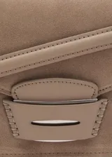 Bolso de hombro tipo baguette confeccionado en piel de serraje color tostado. Presenta un diseño rectangular con solapa frontal decorada con un detalle metálico y pespuntes visibles. Incluye un asa fija y forro interior.