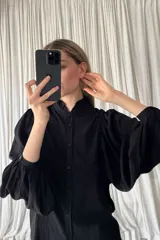Camisa negra de corte clásico con cuello alto tipo mao con volados, cierre frontal de botones y mangas largas abullonadas con puños ajustados.