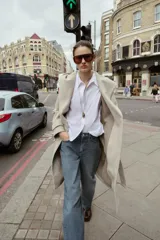 Abrigo de paño negro con capucha, cierre frontal cruzado con botones y cinturón de lazada en el mismo tejido. Presenta manga larga con trabillas en los puños y bolsillos laterales.