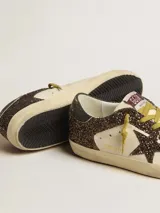 Zapatillas deportivas Golden Goose modelo Super-Star, con cuerpo principal de cuero color crema y detalles de glitter marrón oscuro. Presentan la estrella lateral y el talón en glitter marrón oscuro, cordones amarillos y suela desgastada.