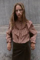 Blusa de manga larga con estampado de cuadros vichy en tonos burdeos y beige. Presenta cuello mao y grandes volantes fruncidos que cruzan el pecho y los hombros. Los puños son elásticos y fruncidos.