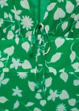 Vestido corto de tejido fluido color verde con estampado floral en tono crema. Presenta escote en pico con cierre de lazo frontal, mangas cortas con vuelo, diseño entallado en la cintura y bajo con volante.
