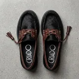 Mocasín de estilo náutico confeccionado en cuero, combinando una base de cuero con textura de pelo en color negro y detalles de cuero con textura croco en color marrón. Presenta cordones decorativos con borlas laterales y suela plana de goma.