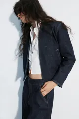 Cazadora bomber de denim color azul oscuro, con escote en pico, cierre frontal cruzado con botones a presión y bajo elástico en los laterales. Cuenta con mangas largas y bolsillos de vivo.