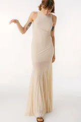 Vestido largo de color beige, con cuello alto y diseño sin mangas. Presenta un corte ajustado en la parte superior y una falda con paneles de tela semitransparente que aportan movimiento y fluidez.