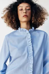 Blusa celeste de popelín, con cuello subido y escote de pico, manga larga abullonada terminada en elástico, y detalles de volantes en el cuello y la botonadura frontal con cierre de botones ocultos.