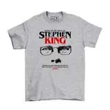 Remera gris de manga corta con estampado frontal temático de Stephen King, que incluye el nombre del autor, un par de anteojos y una frase alusiva al terror.