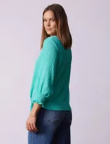 Blusa blanca de tejido texturado con cuello redondo, escote en V con aplique metálico y mangas 3/4 con puños elásticos. El ruedo se ata con un nudo.