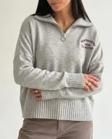 Sweater de tejido bremer color gris melange, con cuello alto y cierre frontal parcial. Presenta mangas largas con puños acanalados y un bordado decorativo en el pecho con la inscripción Montmartre 1974 La Butte.