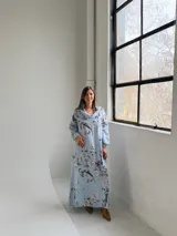 Vestido largo celeste con estampado floral en tonos blancos y grises, de corte holgado, mangas largas y cuello drapeado.