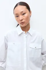 Camisa blanca de manga larga con cuello clásico y botones frontales. Presenta dos bolsillos asimétricos en el pecho, uno con solapa y otro sin.
