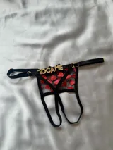 Tanga negra de diseño abierto con tiras regulables, detalles metálicos dorados, aplique de letras en strass con la frase "TOCA ME" en la parte trasera y tejido transparente bordado con corazones rojos.