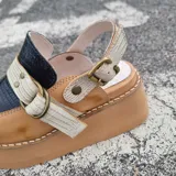 Zueco de cuero color marrón claro, con capellada en cuero negro con textura croco, tiras cruzadas frontales en cuero texturizado claro con hebilla metálica circular y tira ajustable en el talón. Cuenta con suela de plataforma corrida.