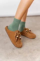 Zueco de cuero gamuzado color tostado, con cordones decorativos adornados con cuentas de madera y piezas envueltas en cuero. Diseño tipo mocasín con costuras visibles en la parte superior y abierto en la parte trasera.