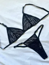 Conjunto de lencería de dos piezas confeccionado en tul transparente con estampado animal print tipo cebra aterciopelado en color negro. Incluye un corpiño triangular sin aro con breteles regulables y una bombacha tipo tanga a juego con detalles de argollas metálicas en los laterales.