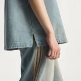 Camisa de jean de manga corta Adidas, confeccionada en 100% algodón, con diseño inspirado en la ropa de trabajo, cierre frontal con broches a presión, cuello camisero, un bolsillo en el pecho con solapa y las clásicas tres tiras de la marca en las mangas.
