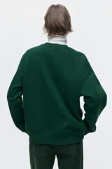 Buzo de cuello redondo en color verde, con diseño gráfico estampado en blanco en la parte frontal. Presenta hombros caídos, corte holgado y terminaciones acanaladas en cuello, puños y ruedo.