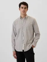 Camisa de manga larga para hombre, color gris claro con finas rayas verticales blancas. Presenta cuello abotonado (button-down) y un bolsillo de parche en el pecho izquierdo. Se lleva abierta sobre una camiseta blanca.