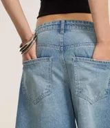Bermuda tipo jorts de tiro medio, confeccionada en denim de algodón color azul claro. Presenta un corte holgado que llega por debajo de la rodilla, con trabillas en la cintura, bolsillos laterales y traseros, y un detalle de cordón decorativo en la parte frontal.