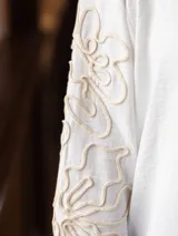 Camisa de color beige con cuello clásico y cierre frontal de botones. Presenta un diseño de bordado lineal en color blanco que recorre las mangas largas.