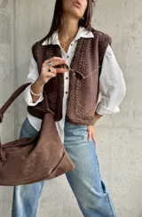 Chaleco de gamuza color chocolate con interior y bordes de corderito sintético. Presenta diseño con tachas metálicas decorativas en el frente y los hombros.