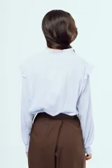 Camisa celeste de manga larga con sutiles rayas verticales, cuello alto mao fruncido y cierre frontal con botones. Presenta volados decorativos en los hombros y el frente, y puños elásticos fruncidos.
