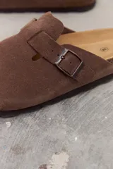 Zueco de gamuza color marrón con diseño de punta cerrada, hebilla metálica lateral ajustable y plantilla de corcho.