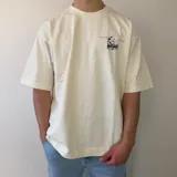 Remera oversize de algodón pesado en color blanco, con cuello redondo y moldería amplia. En la espalda cuenta con un gran parche rectangular estampado con una fotografía en blanco y negro de aves volando sobre el agua y texto tipográfico al margen.