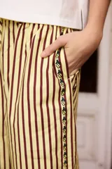 Pantalón de corte recto y tiro alto, confeccionado en tela de algodón con diseño de rayas verticales en tonos oscuros. Presenta una banda lateral decorativa con pequeños detalles bordados en color amarillo.
