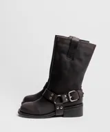 Bota de caña media confeccionada en cuero vacuno color marrón oscuro, con diseño de estilo biker. Presenta una tira decorativa con hebilla metálica y estribo desmontable en la zona del tobillo, taco cuadrado de madera y suela de caucho.