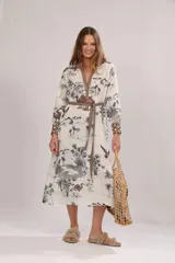 Túnica midi color blanco de manga larga, con escote en V profundo y lazo ajustable en la cintura. Presenta un estampado de paisaje desértico con palmeras y camellos, además de detalles geométricos en el escote, puños y cinturón.