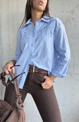 Camisa celeste de manga larga con cuello clásico, botones frontales y bordados florales en el pecho.
