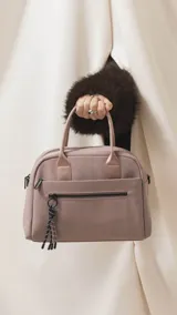Cartera de mano color beige, confeccionada en tela impermeable, con doble asa corta y correa larga ajustable y desmontable. Presenta cierre principal, bolsillo frontal con cierre y detalle de colgante decorativo.