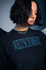 Remera negra de corte holgado con estampa frontal en color turquesa que forma la palabra Weltschmerz con un diseño de puntos.