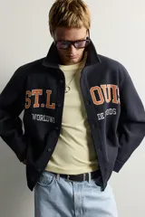 Sobrecamisa de ajuste relajado en color azul marino, con cuello de solapa, manga larga con puño abotonado y cierre frontal con botones. Presenta bordados contrastantes en el frente con la inscripción "St. Louis Worldwide Goods" en tonos naranja y blanco.