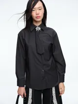 Camisa de corte clásico en color gris oscuro, con cuello camisero, mangas largas con puños abotonados y cierre frontal con botones. Incluye una corbata a tono con un broche decorativo en forma de flor metálica en el cuello.