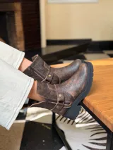 Bota corta de cuero vacuno en color chocolate con efecto desgastado. Presenta una suela track de goma negra, detalle de pulsera desmontable con tachas metálicas y cierre lateral.