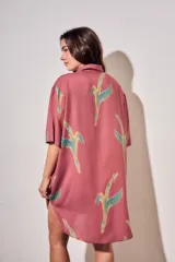 Vestido camisero corto de color rosado con estampado de aves abstractas en tonos verdes y azules. Cuenta con cuello camisero, cierre frontal con botones, mangas cortas y un bolsillo aplicado en el pecho.