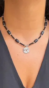 Collar corto tipo gargantilla compuesto por cuentas negras facetadas y cuentas cilíndricas negras lisas, intercaladas con cuentas espaciadoras plateadas. Incluye un dije central plateado con diseño de flor o sol calado.